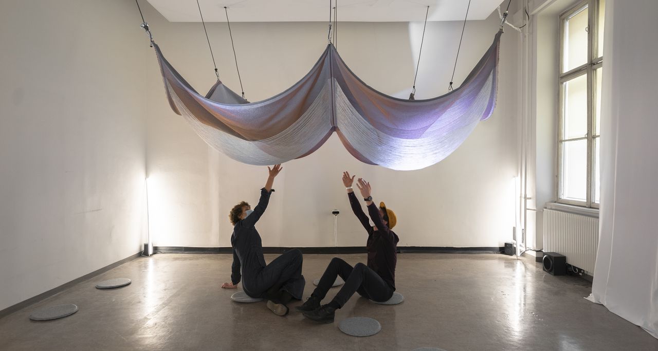 Two people sitting on the floor, the woven textile installation on the ceiling is slowly coming closer.